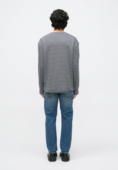 Kékfehér hosszú ujjú csíkos póló, laza szabással. Világoskék egyenes szárú farmerrel és fekete lábbelivel párosítva.