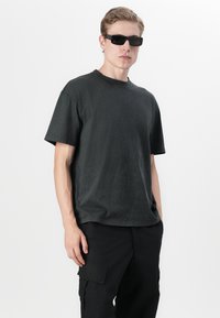 Νεαρός άνδρας με μαύρα γυαλιά ηλίου, μαύρο κοντομάνικο μπλουζάκι και μαύρο παντελόνι cargo, που στέκεται μπροστά σε λευκό απλό φόντο.