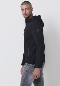 Jachetă neagră din softshell cu glugă, dotată cu buzunare cu fermoar și un logo subtil pe mânecă. Textură netedă și design ajustat.