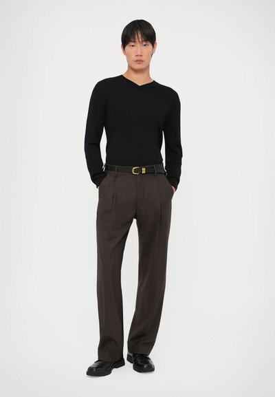 Suéter preto canelado de manga longa com decote em V, combinado com calças marrons plissadas e um cinto preto; os sapatos são pretos, de material durável.