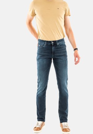 Vaqueros de mezclilla azul oscuro de corte slim, con cinco bolsillos y detalles de costura sutiles, combinados con una camiseta de cuello redondo beige y zapatillas marrones.