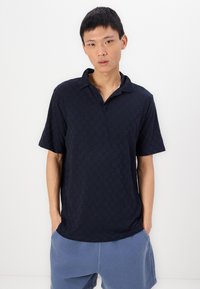 Μαρινί μπλουζάκι πόλο με υφασμένη καρό σχεδίαση, κοντά μανίκια και άνετη εφαρμογή. Διαθέτει γιακά και διακριτική πατιλέτα στο λαιμό.