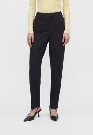 Un mannequin porte un pantalon noir à jambes droites, un haut jaune pâle et des escarpins noirs à bride arrière, sur un fond clair uni.