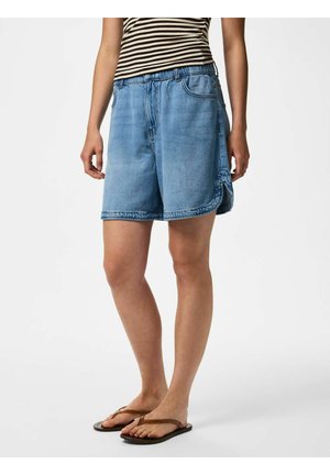 Person iført lyseblå denimshorts, en sort-hvid stribet ærmeløs top og brune flip-flops, der står på en hvid baggrund.