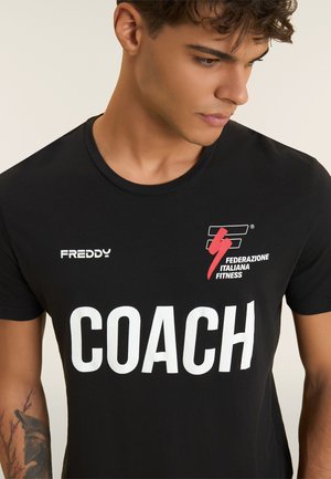 Joven mirando hacia abajo, vistiendo una camiseta negra con "COACH" en blanco, el logo de "FREDDY" y el emblema de la "Federazione Italiana Fitness" en el pecho.
