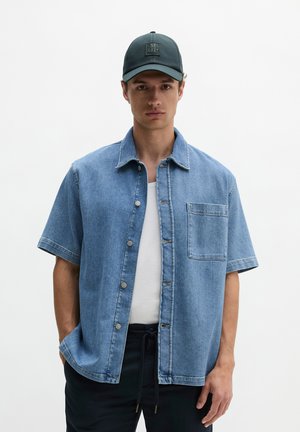 Junger Mann trägt ein blaues kurzärmeliges Jeanshemd über einem weißen Tanktop und eine dunkle Kappe mit dem "M OP"-Logo vor einfarbigem Hintergrund.