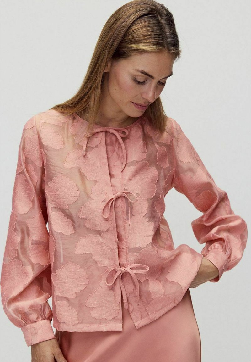 Bluzka w odcieniu brzoskwiniowym, wykonana z przezroczystego materiału, zdobiona wyszywanymi wzorami liści. Posiada zapięcie na węzeł z przodu oraz balonowe rękawy z elastycznymi mankietami.