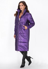 Cappotto lungo imbottito viola con colletto alto, chiusura con zip e tasche laterali. Texture liscia e lucida, abbinato a stivali neri fino al ginocchio.