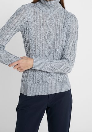 Jasnoszary golf z dzianiny warkoczowej noszony z ciemnymi spodniami, pokazujący teksturowane wzory ściegów oraz osobę poprawiającą rękaw.