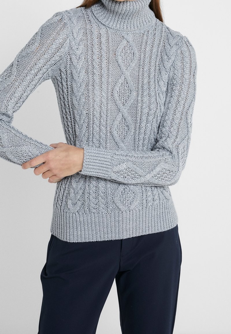 Pull à col roulé gris clair en maille torsadée porté avec un pantalon sombre, montrant des motifs de points texturés et une personne ajustant la manche.