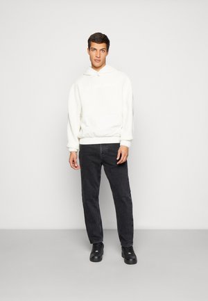 Hvit fleece-hettegenser med frontlomme og logo, kombinert med svarte denimjeans og svarte joggesko, stående mot en ensfarget bakgrunn.