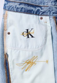 Εσωτερικό τσέπης από ανοιχτό denim με κεντημένο μαύρο και κίτρινο λογότυπο «Jungkook JK» και κίτρινη υπογραφή με καλλιγραφική γραφή, ραμμένο με αντιθετικές ραφές και φιλέτα.