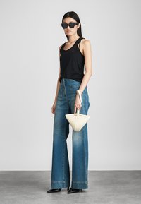 Melnā topiņa bez piedurknēm, augstās jostas zilās džinsās, melni saulesbrilles, tur rokā krēmkrāsas trīsstūra soma. Gluda denim auduma tekstūra, plata kāju dizains.