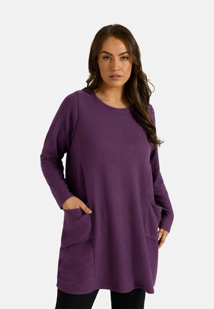 Femme portant une tunique violette ample à manches longues avec des poches, debout devant un fond clair uni.