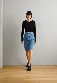 Čierny top s dlhými rukávmi, sukňa zo svetlomodrej rifloviny s vysokým pásom a čierne topánky. Modelka kráča po drevenej podlahe pred šedou stenou.