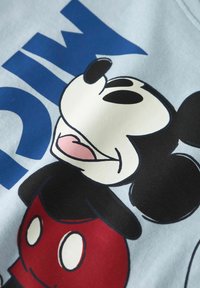 Myšiak Mickey v červených šortkách a čiernych ušiach sa usmieva s textom "TERAZ" v tučných modrých písmenách na svetlomodrom pozadí.