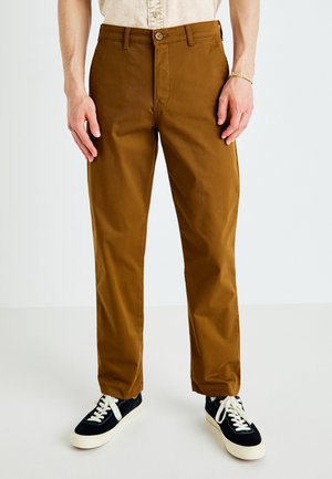 Chino in cotone marrone con taglio dritto, dotato di chiusura a bottone e tasche laterali, abbinato a sneakers bianche e nere.