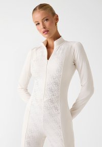 Combinaison blanche à manches longues avec un col montant et une fermeture éclair, présentant un motif en losanges texturé sur le devant. Matière lisse et extensible.