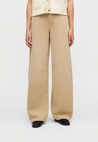 Άτομο που φοράει μπεζ παντελόνι με φαρδιά μπατζάκια, μαύρα παπούτσια και μπεζ αμάνικη μπλούζα με κουμπιά, στέκεται μπροστά σε ένα απλό φόντο.