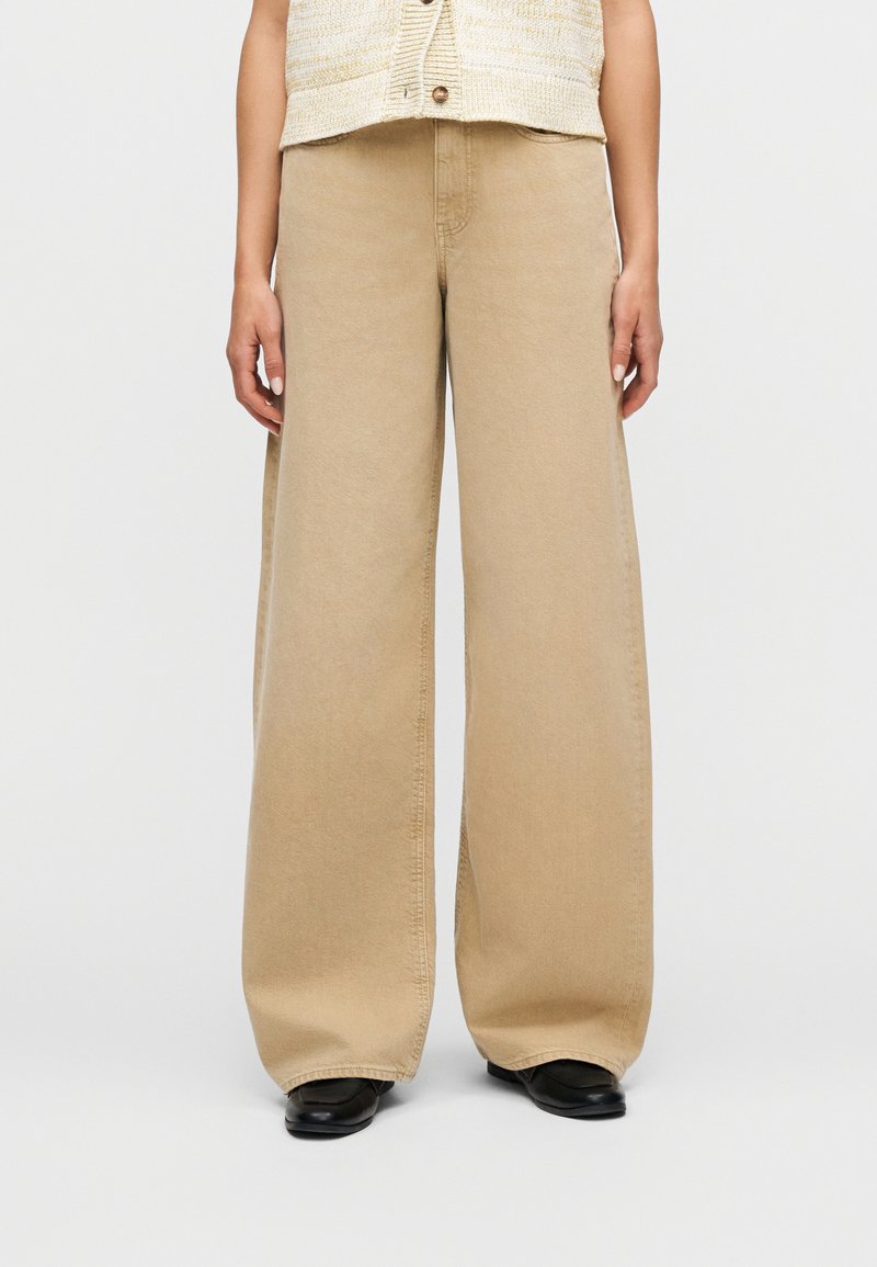 Άτομο που φοράει μπεζ παντελόνι με φαρδιά μπατζάκια, μαύρα παπούτσια και μπεζ αμάνικη μπλούζα με κουμπιά, στέκεται μπροστά σε ένα απλό φόντο.