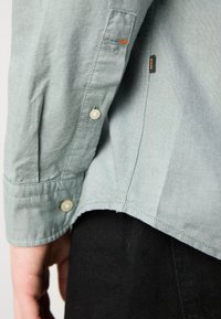 Lichtgroen, lang mouwenshirt met een knoopmanchet, met een zijsplit en een oranje accentsteek. Draagt over zwarte broek.