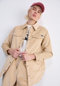 Beige cordfløyelsjakke med to frontlommer og trykknapper, kombinert med en lysfarget t-skjorte og matchende bukser med et mønstret snor.