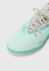 Světle tyrkysová a béžová sportovní bota Nike s texturovaným svrškem, béžové tkaničky uvázané do mašle a malý oranžový znak Nike na boku.