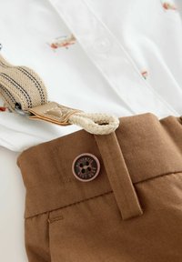 Light brown trousers with a wide waistband, featuring a dark brown button labeled “AUTHENTIC NEXT DUDE” and a beige striped suspender strap.