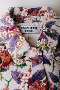 Μπουφάν με μοτίβο λουλουδιών σε μπεζ βάση, με μοβ, πράσινες και κόκκινες αποχρώσεις. Η ετικέτα γράφει "COLOURFUL REBEL", μέγεθος S. Υφασμένο ύφασμα με υφή.