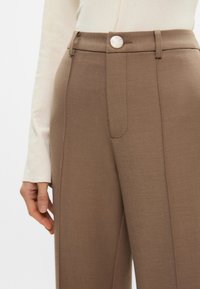 Καφέ ραμμένα παντελόνια με λεία υφή, που διαθέτουν πτυχές μπροστά, πλαϊνές τσέπες και γυαλιστερό μεταλλικό κλείσιμο.