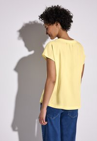 Blusa gialla a maniche corte con dettaglio plissettato sul retro, abbinata a jeans in denim blu, mostrata da un angolo laterale e posteriore.