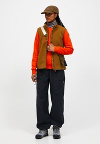 Braune Fleece-Weste über einem orangefarbenen Sweatshirt, kombiniert mit schwarzen Cargo-Hosen. Ausgestattet mit grauen Wanderschuhen und einer braunen Kappe. Strukturiertes, funktionales Design.