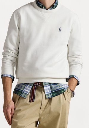 Mężczyzna ubrany w białą bluzę na zielono-niebieskiej koszuli w kratę, beżowe spodnie i paskiem z tkaniny w paski, ręce w kieszeniach, na nadgarstku brązowy zegarek.