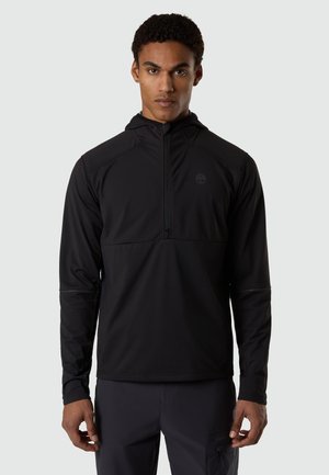 Homme portant un pull noir ajusté à manches longues avec une demi-fermeture éclair et une capuche, debout devant un fond clair uni.