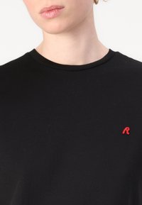 Svart bomull t-skjorte med rund hals, med en liten rød brodert "R" på venstre bryst. Glatt tekstur, tettsittende design.