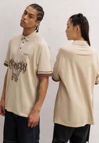 Beige polo skjorte med brune detaljer, med et sommerfugl grafikk. Laget av myk bomull, har den en avslappet passform og kortermer.