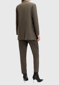 Femme portant une veste et un pantalon de costume vert olive sur mesure avec des bottines noires pointues à talons hauts, vue de dos.