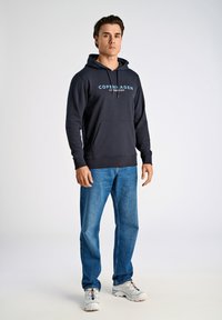Felpa navy con tasca a marsupio e coulisse, con il testo "COPENHAGEN" di colore azzurro. Abbinata a jeans azzurri e sneakers bianche.
