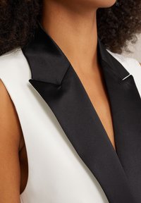 Kvinna med en ärmlös vit blazer med bred svart satinrevers och lockigt mörkt hår synligt.