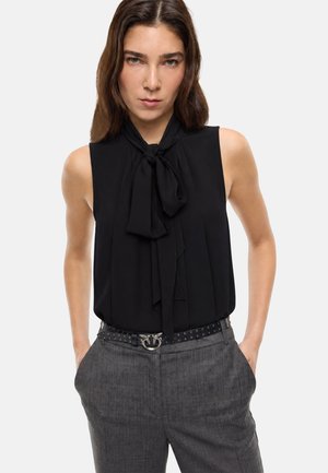Donna che indossa una camicetta nera senza maniche con colletto annodato a fiocco e pantaloni grigi a vita alta con cintura nera con borchie.