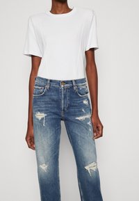 Camiseta blanca de manga corta, con escote redondo, combinada con jeans anchos de mezclilla azul desgastados que presentan desgarros y áreas desteñidas, con cierre de botón y cremallera.