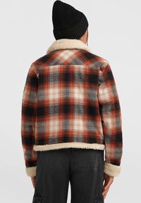 Persoon met een rood, zwart en wit geruite jas met een crèmekleurige fleece rand en een zwarte gebreide beanie, met de rug naar de camera gekeerd.