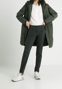 Abrigo acolchado verde oliva sobre una camisa blanca de botones, combinado con pantalones de cuadros negros y zapatillas blancas con acentos grises.
