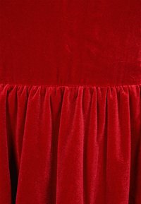 Piros bársonyos anyag sima textúrával és ráncolt díszítéssel az alján, amely gazdag, mély színt és diszkrét fényt mutat.