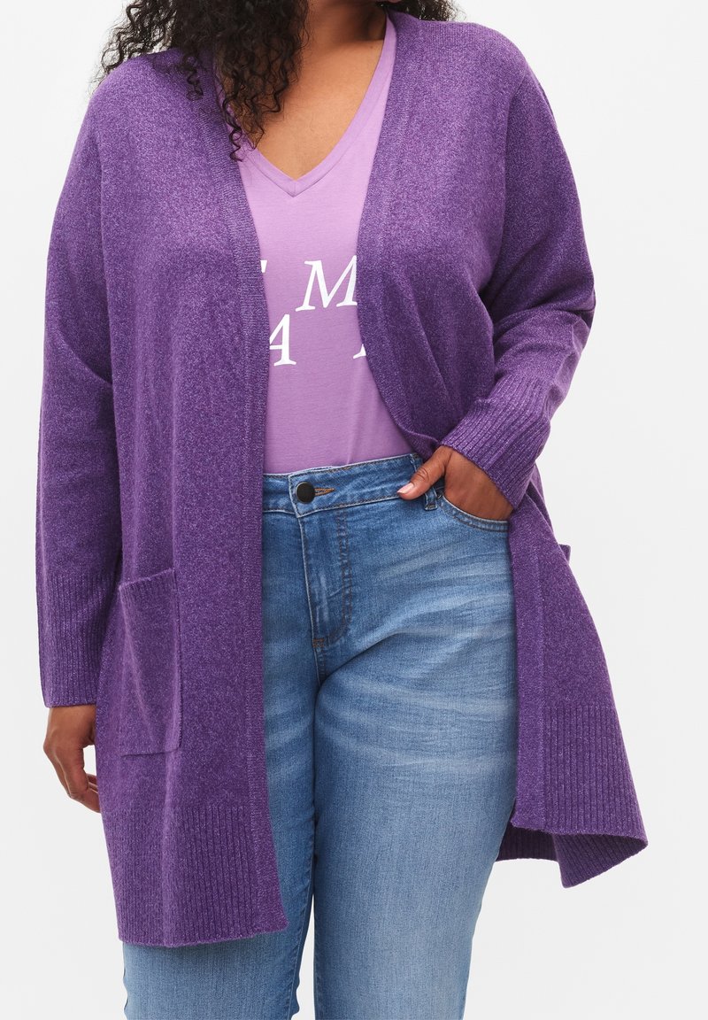 Femme portant un cardigan violet ouvert sur un T-shirt lavande à col en V avec un texte blanc, et un jean denim bleu clair, main dans la poche.