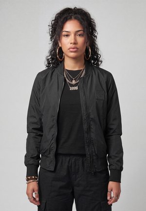 Young woman with curly hair wearing large hoop earrings, layered gold necklaces, a black jacket, black shirt, and black pants, standing against gray background.