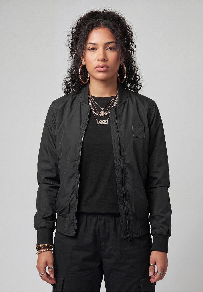 Young woman with curly hair wearing large hoop earrings, layered gold necklaces, a black jacket, black shirt, and black pants, standing against gray background.