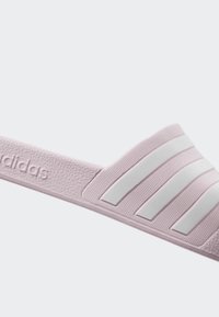 Ružičaste adidas natikače s teksturiranim trakama, s tri bijele paralelne pruge i ugraviranim logom sa strane. Ravni dizajn, lagane.
