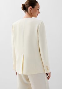 Blazer color crema con un tejido texturizado, corte cuadrado en la parte posterior, una abertura y puños con tres botones. Llevado con pantalones a juego.