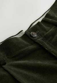 Olijfgroene corduroy broeken met een ritssluiting en een decoratieve knop. De stof heeft een ribbeltextuur en een gladde binnenvoering.